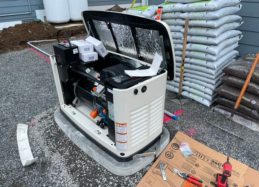 Generator installation with opened cover, tools, and landscaping materials in background.